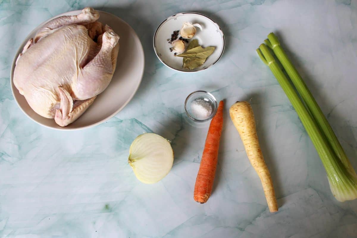 Recipe ingredients on a light blue-marble backgrounds: a whole thawed bird, half of the onion, a carrot, a parsnip, celery, salt, garlic, whole peppercorn and bay leaves.