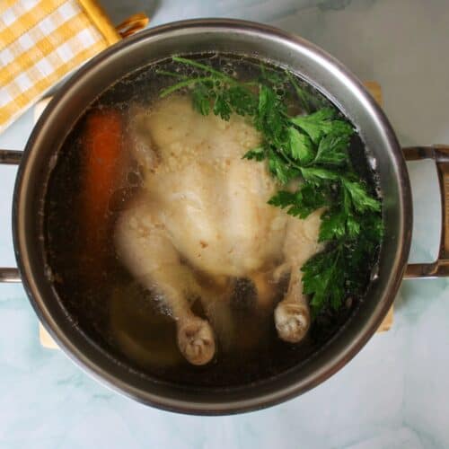 Overhead view of large stainless steel pot with whole chicken, broth, carrot and green parsley in it.