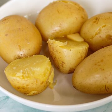 Cooked whole steamed potatoes on a white plate. One potato is cut in half and there is a small piece of butter on top..