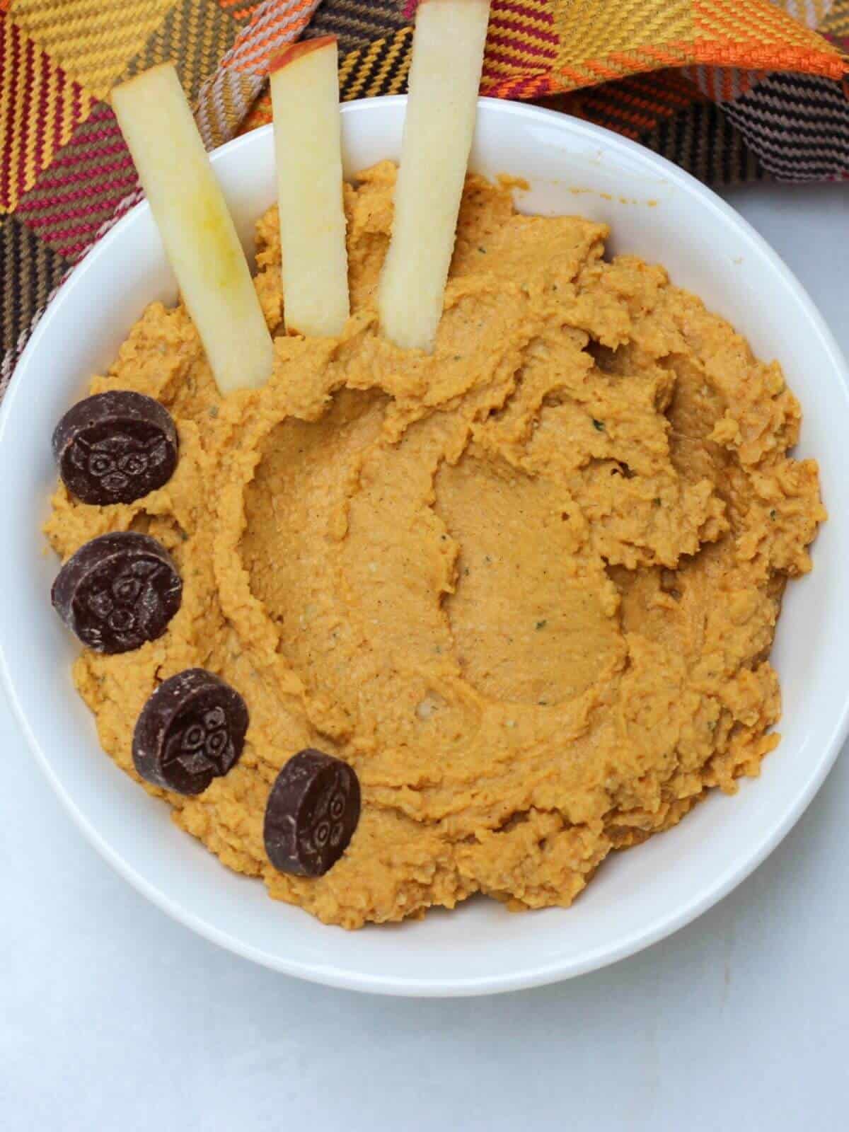 A bowl with orange dip topped with some chocolate and three apple sticks.