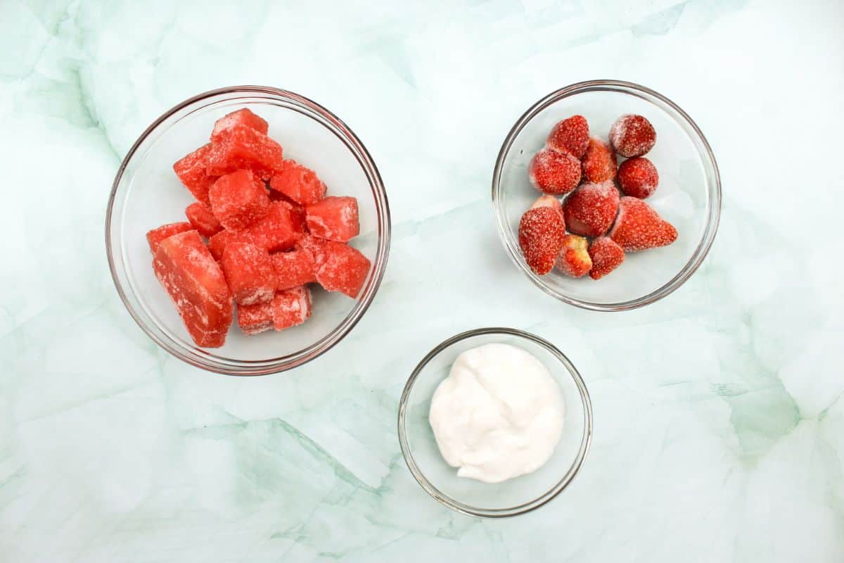 Ingredients to make a watermelon smoothie bowl arranged on a white and green marble background. There are frozen watermelon cubes, frozen strawberries and yogurt.