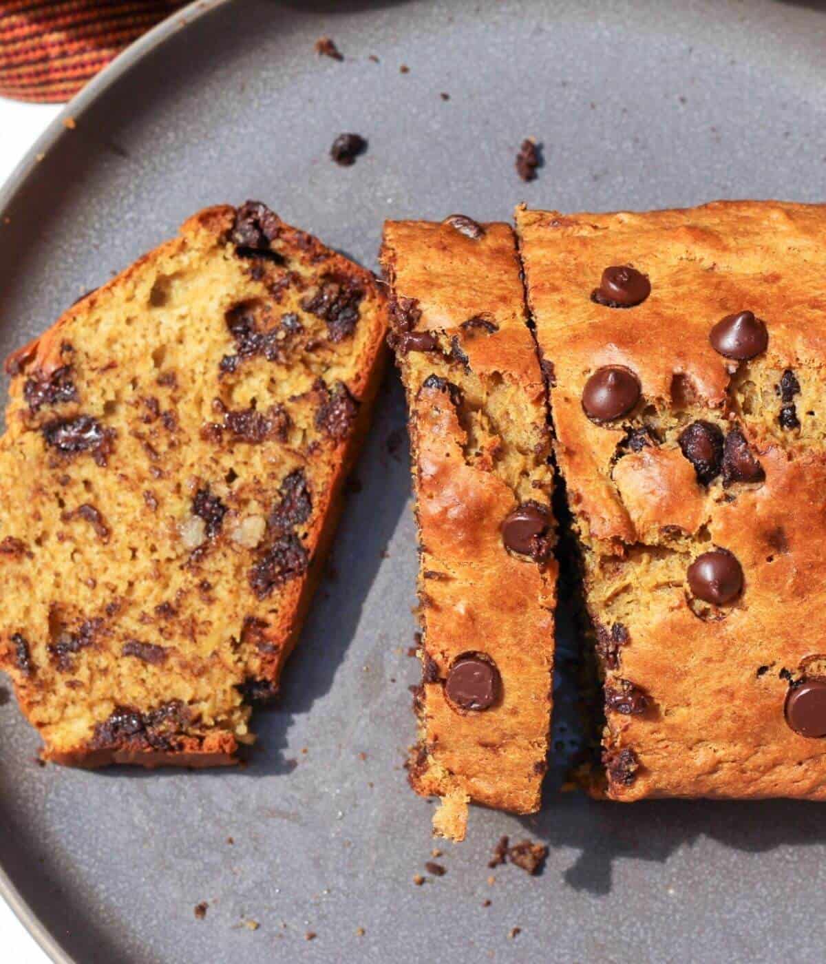 A loaf with chocolate chips on a dark-grey plate. The half of bread is sliced and one slice is laying flat.