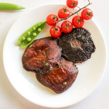 Three cooked large portobello mushrooms on a white plate. There is a vine with cherry tomatoes and an open pod of snap pea on top.