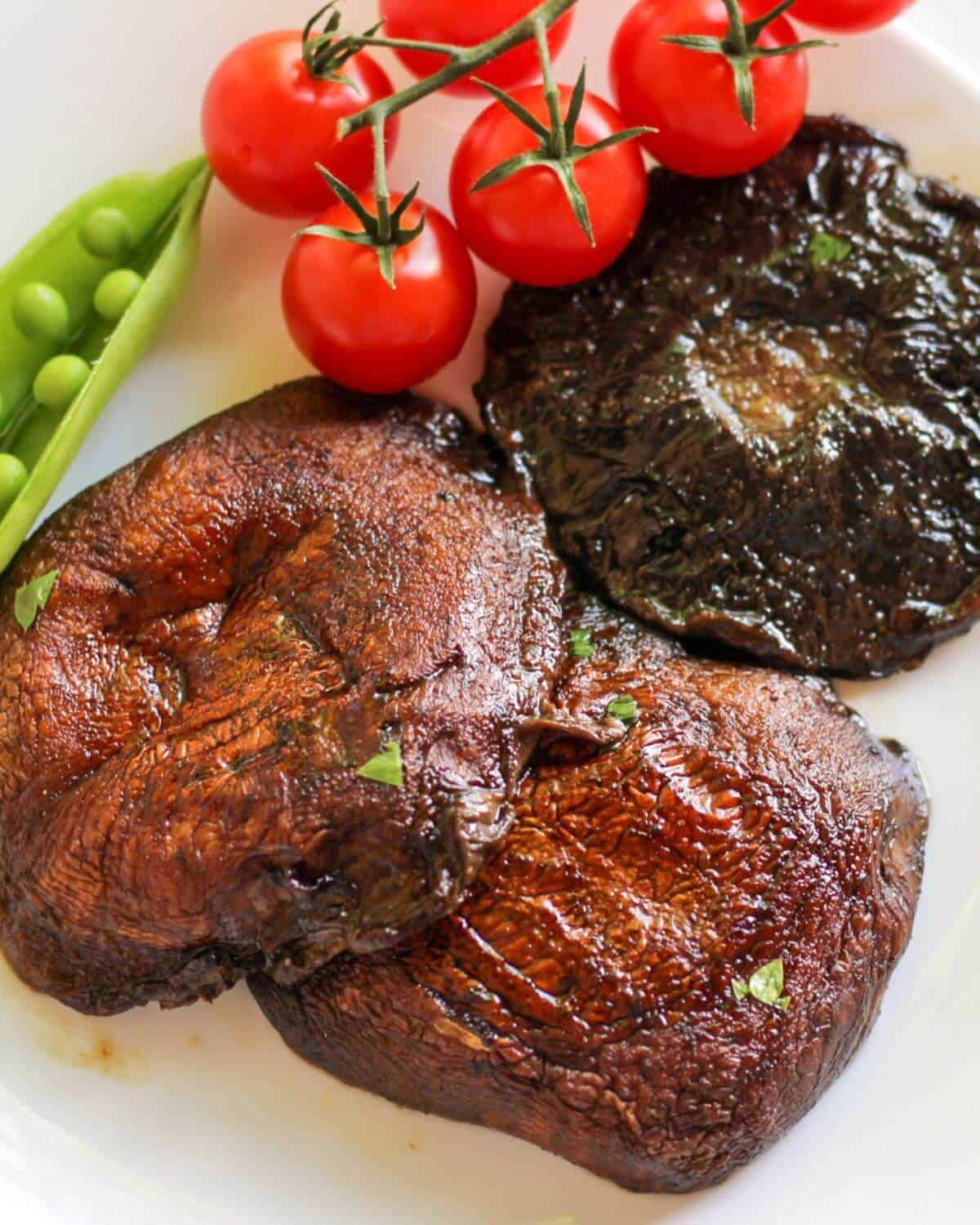 Three cooked mushroom caps in a white place with some cherry tomatoes and open pod of snap pea on a side.