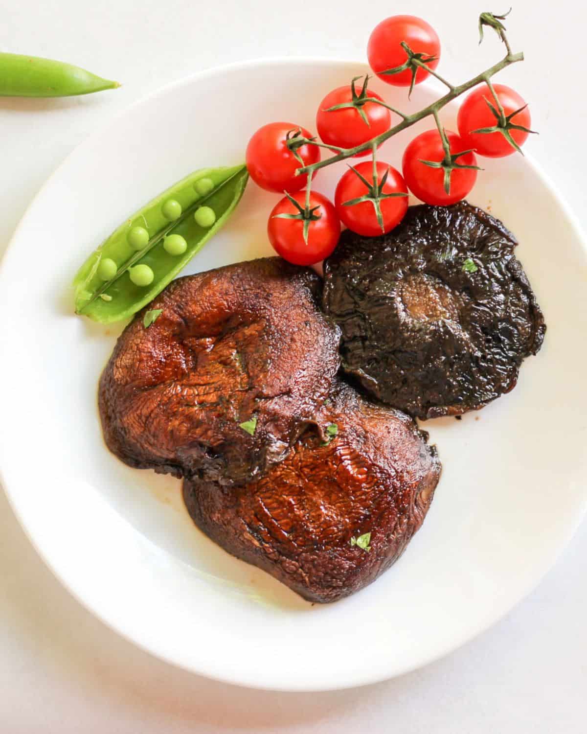 Overhead view of a white plate with three cooked large mushrooms. There is cherry tomatoes on a vine and an open green pea pod on top of the plate.