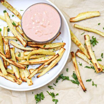 White plate with golden fries and a small bowl of a pink dipping sauce. There is chopped green herbs on top of veggies and on the surface around the plate.
