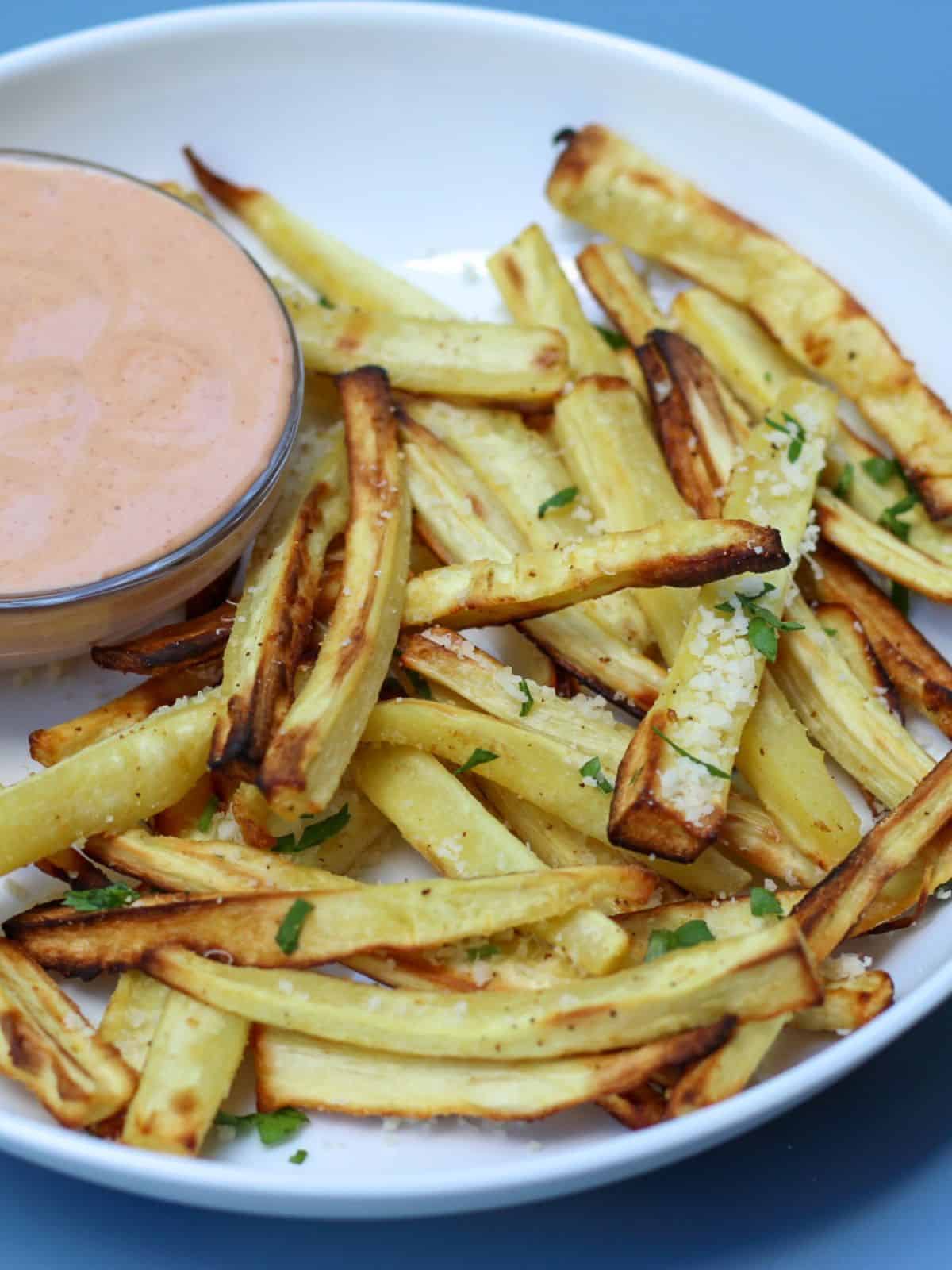 Air fryer parsnip fries served on a white plate. There is visible small bowl with a dipping sauce on a side.