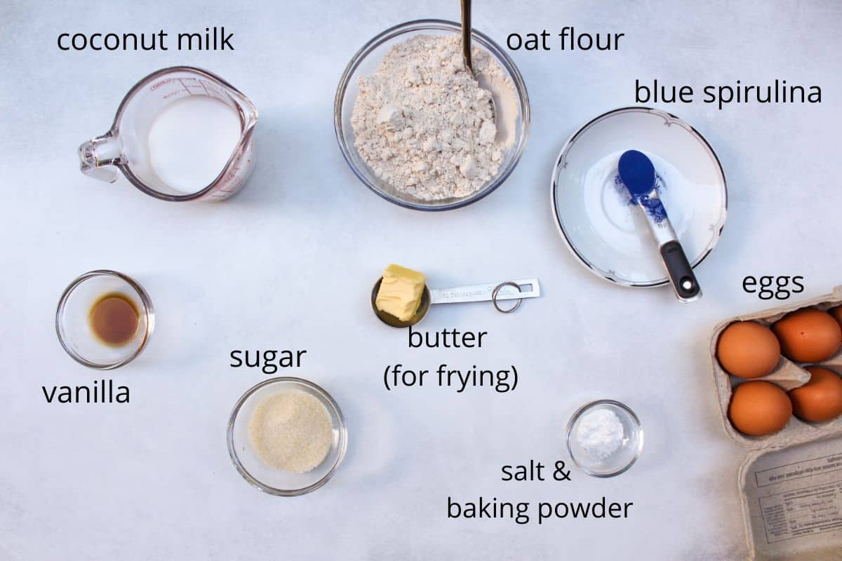 Labeled recipe ingredients on a white background. There is milk, flour, blue spirulina, vanilla, sugar, butter, baking powder with salt, and some eggs.
