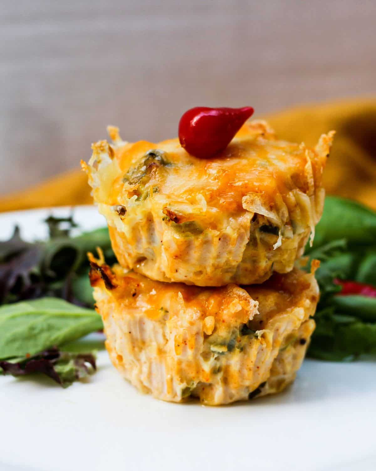 Two savory golden muffins sracked on top of each other. There is some greens on the background and a small bright red pepper on top of muffins.