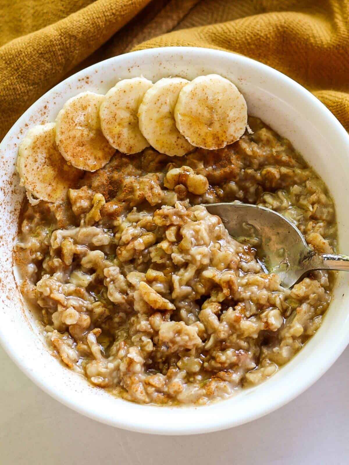 Overhead shot of a white bowl filled with cooked oatmeal. It is sprinkled with brown sliced and has sliced bananas on a side. There is a spoon dipped in the porridge.