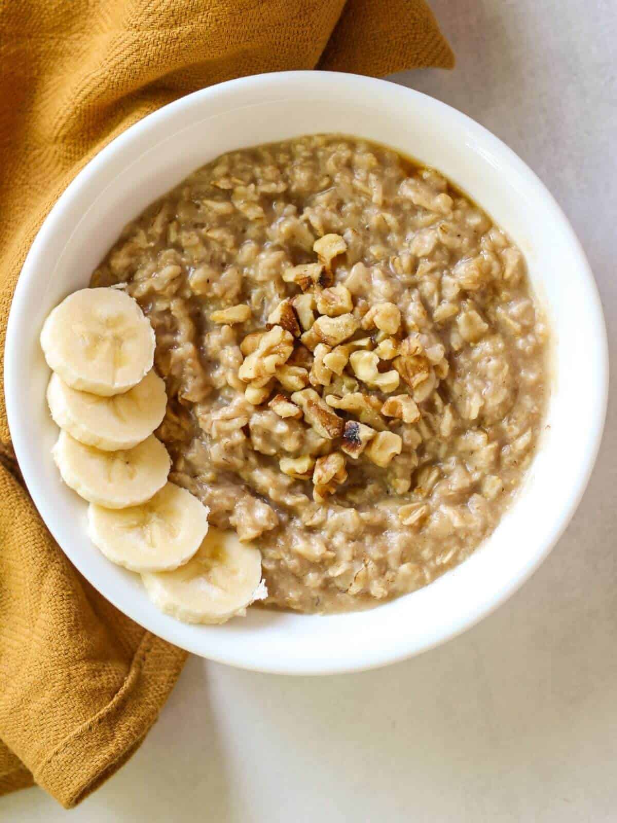 Overhead shot of a white bowl with cooked oats topped with walnuts and slices bananas.