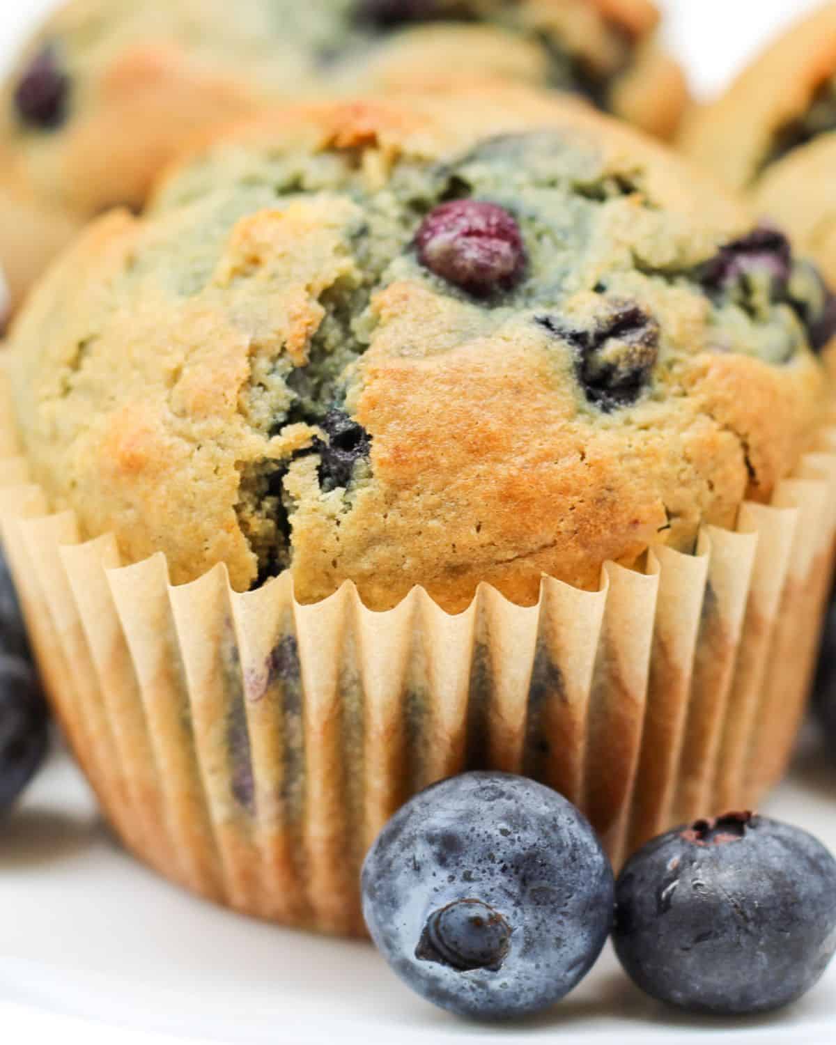 Close-up shot of large golden muffin. It is in a parchment paper liner and there are few fresh blueberries next to it.