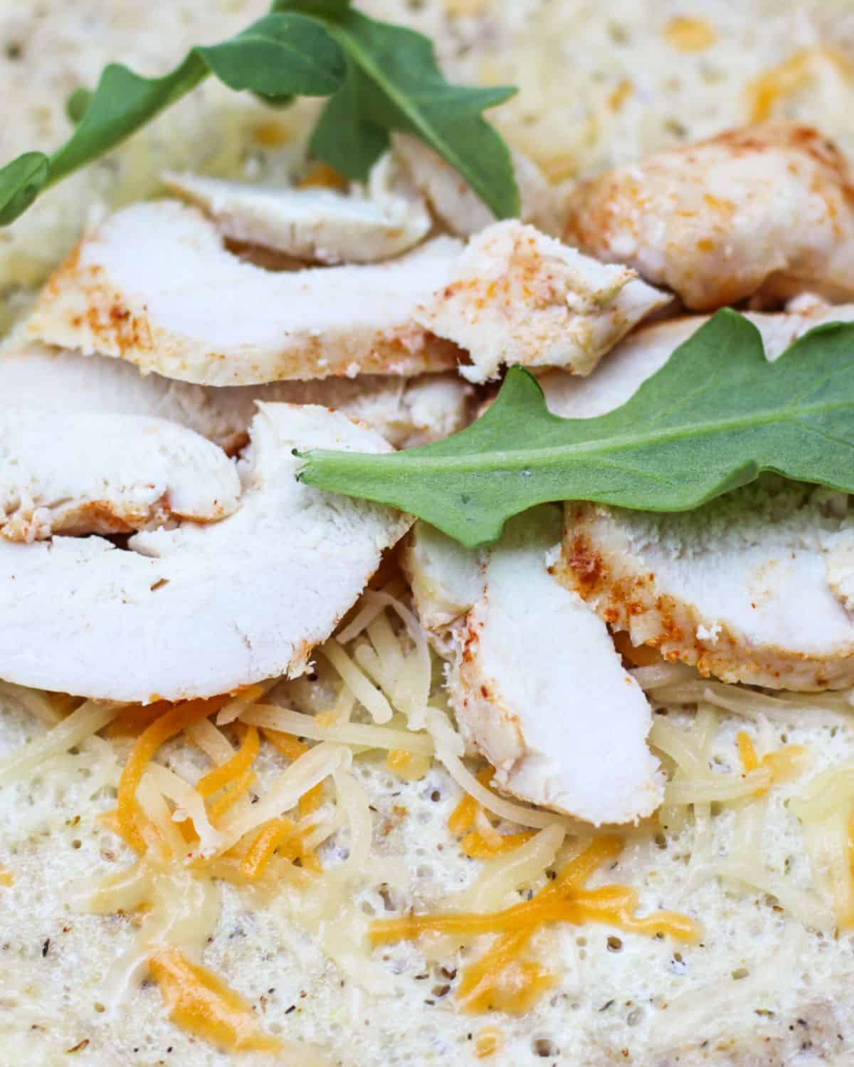 Sliced cooked chicken breast with cheese and few arugula leaves on top of a flat wrap.