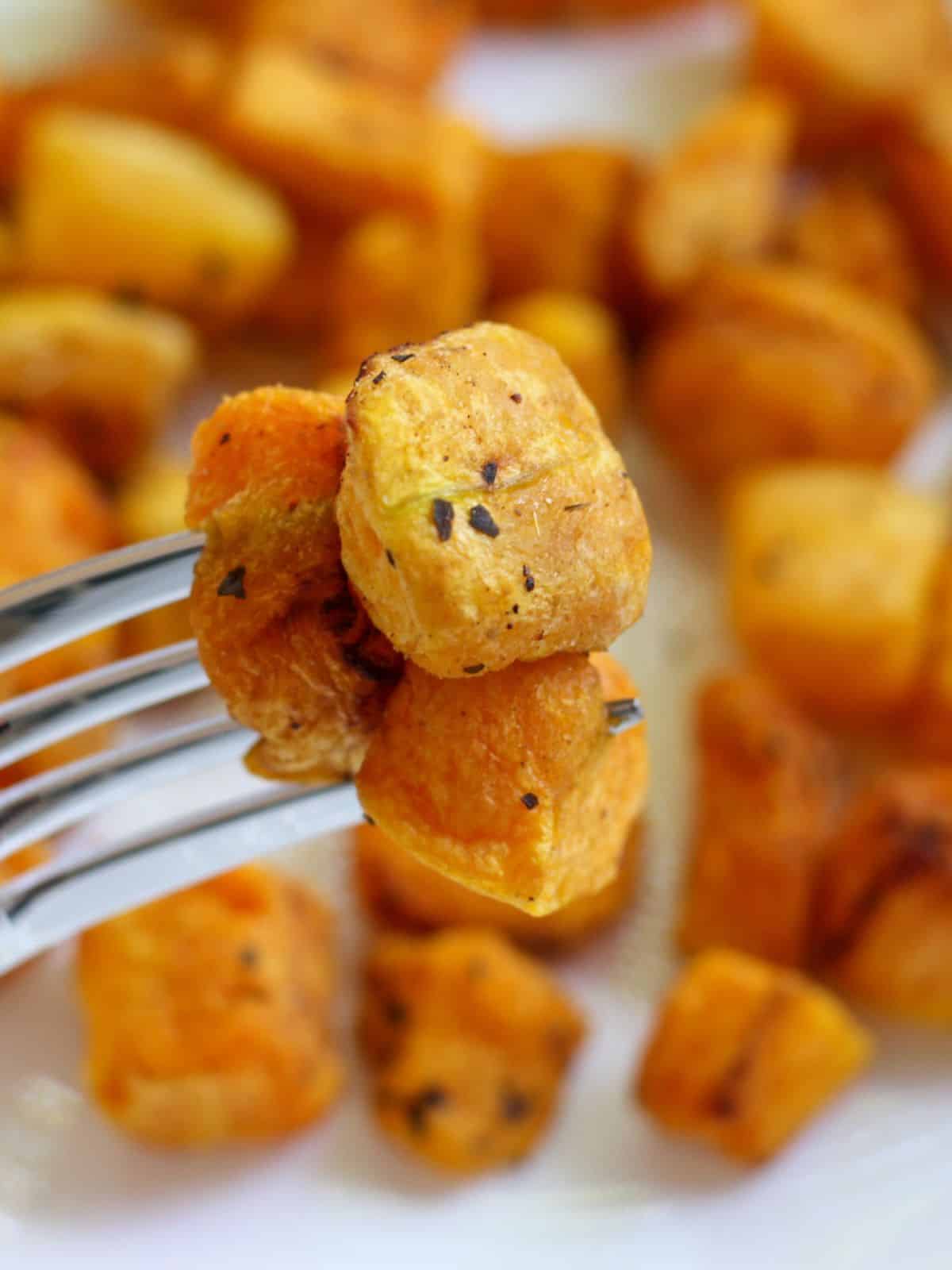 A close-up shot of a fork with 3 cubes of roasted squash and a blurred background with the rest of the vegetables.