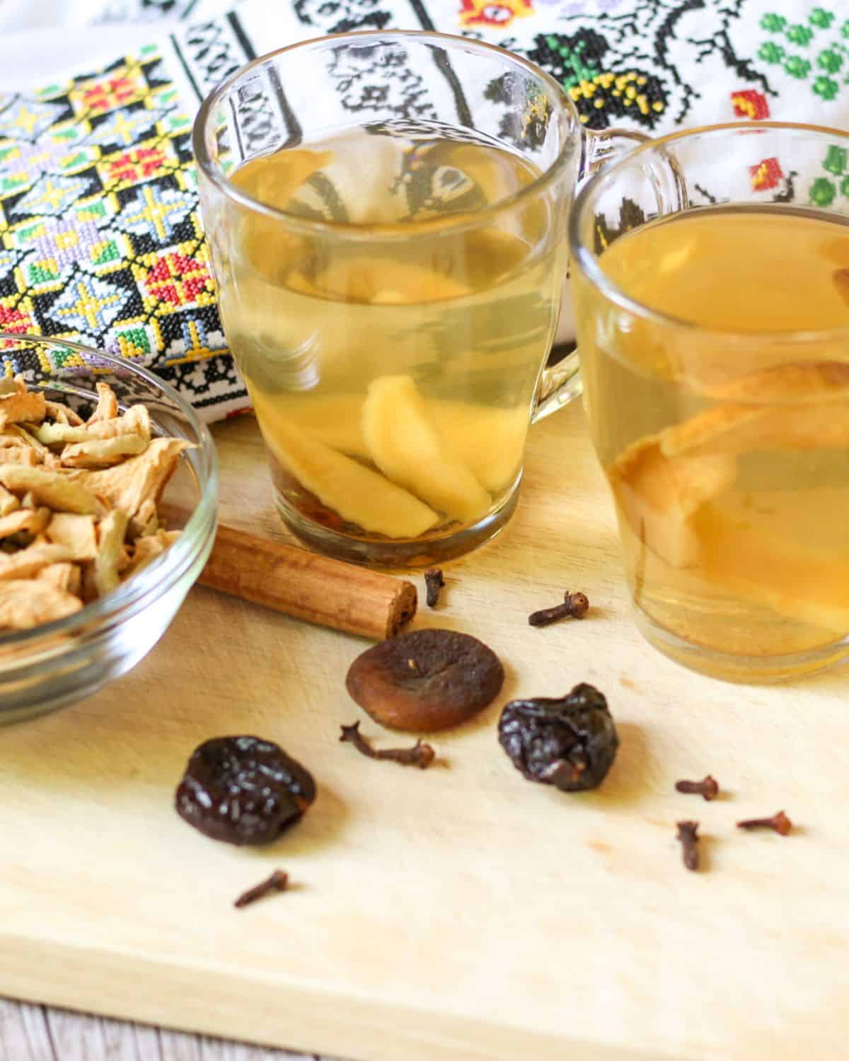 Two clear cups with a stewed fruit drink are placed on a wooden table. There is a cinnamon stick, some dried fruits, and whole cloves next to the cups. The background has a towel with Ukrainian embroidery.