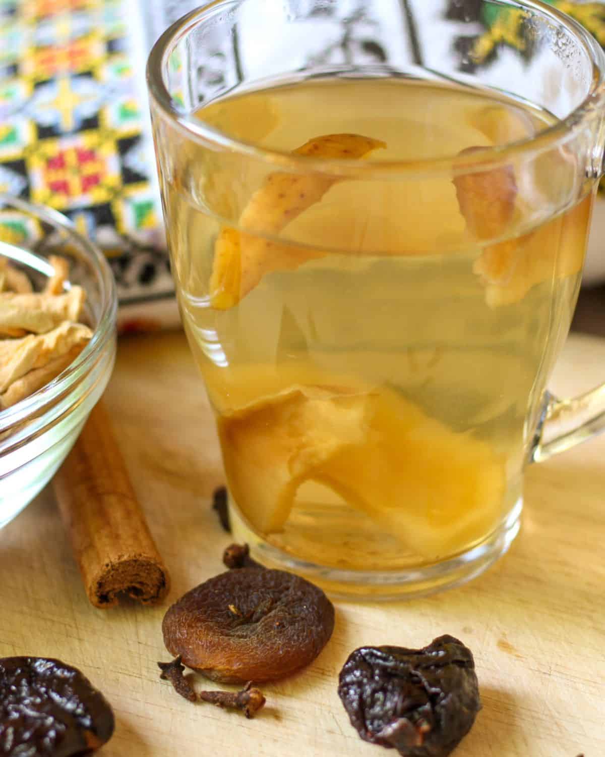 A glass cup filled with a yellow drink with some slices of fruits inside. There is a cinnamon stick in a table and few dried fruits. Ukrainian embroidery on the background.