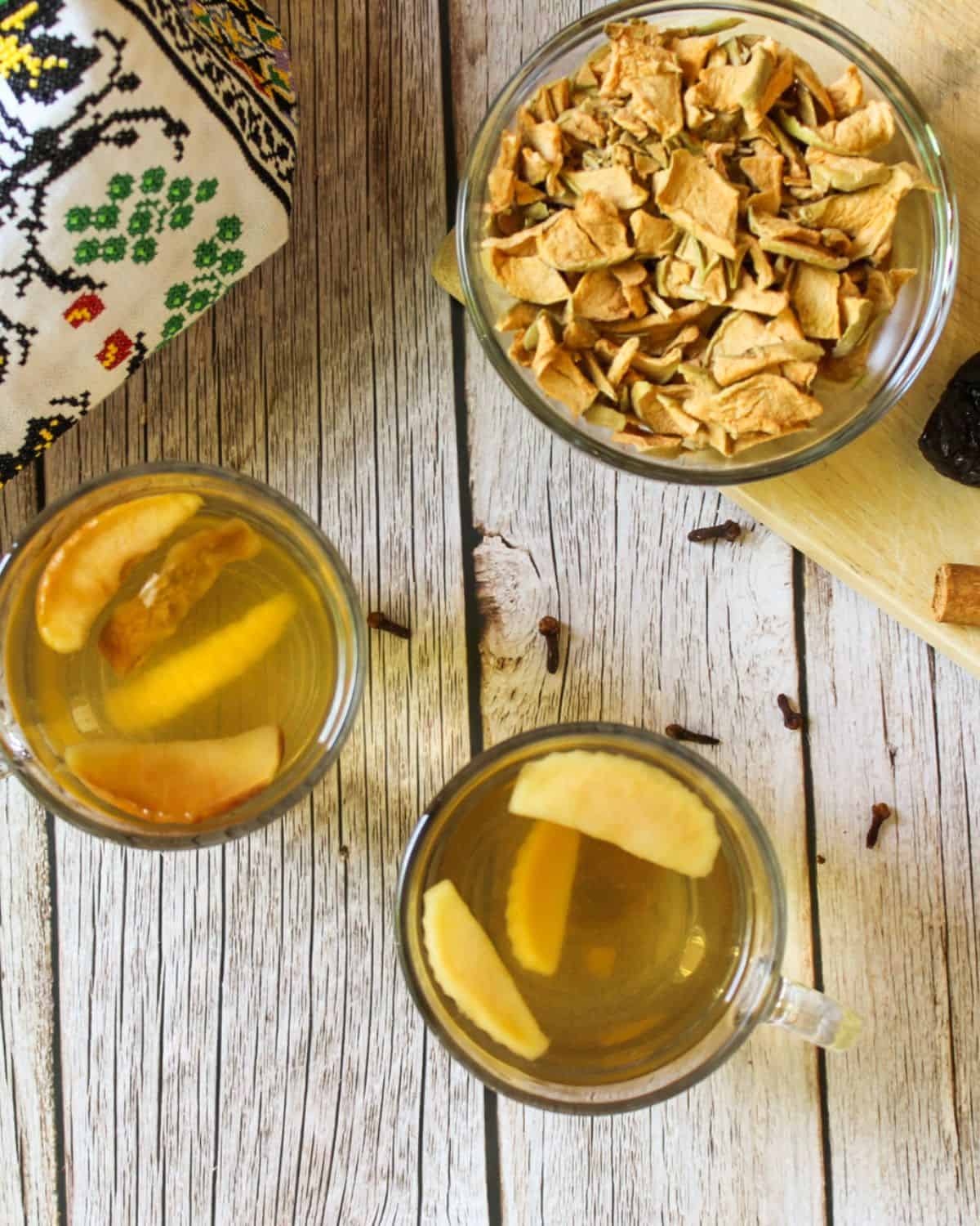 Overhead view of two cups filled with yellow drink and slices of fruits. A glass bowl with some dried apples and a corner of the cloth with Ukrainian embroidery on a wooden background.