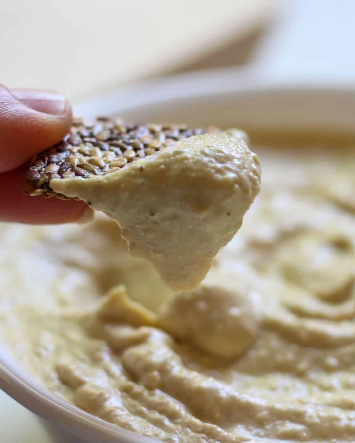 Close-up shot of a dark seed cracker dipped in hummus and held over the  blurred bowl in the background.