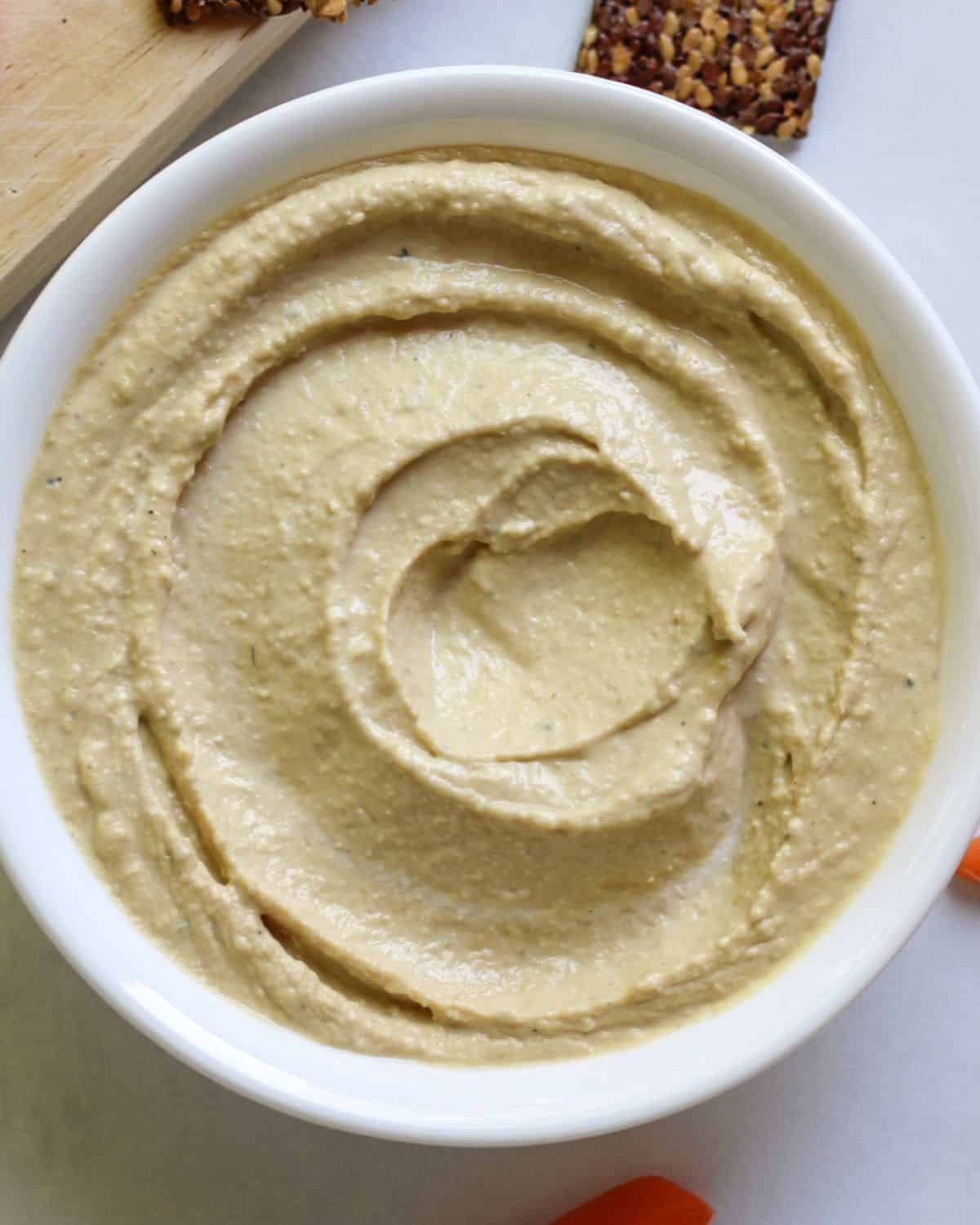 Overhead view of a white bowl with a thick dip that is swirled on top. There is a flex seed cracker behind the bowl.