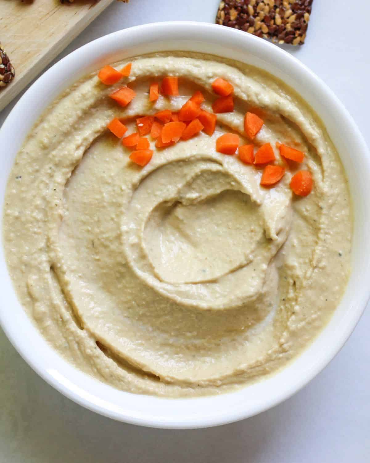 A creamy hummus swirled in a white bowl. There is diced orange carrots on the top side.  