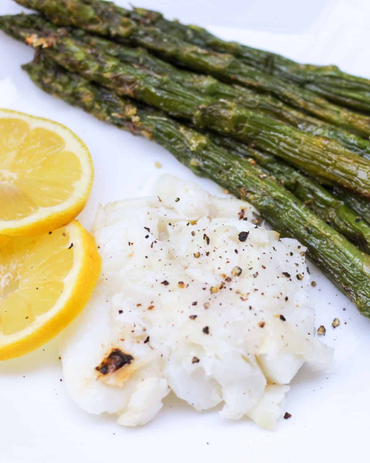 A plate with white fish meat, roasted asparagus and fresh sliced lemon slices on a side.