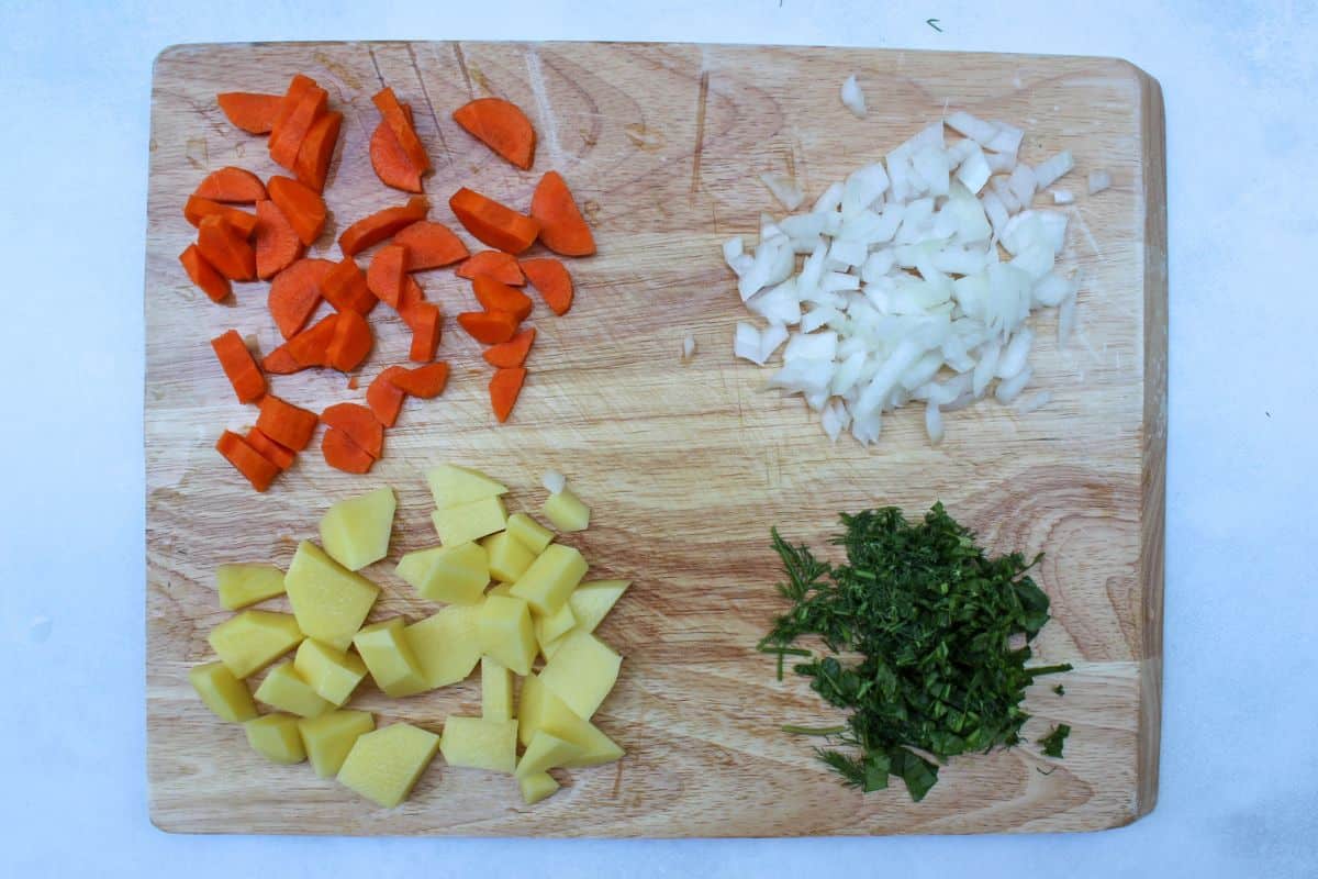 A cutting board with chopped carrots, potatoes, onion, and green herbs.