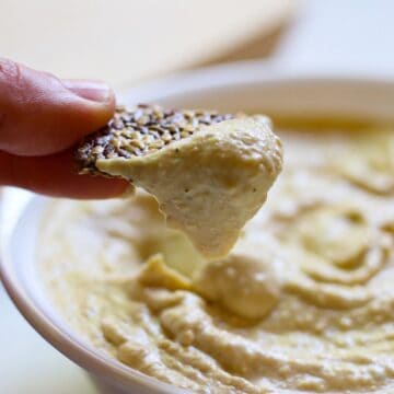 Cracker dipped in high-protein hummus held over the white bowl with hummus.