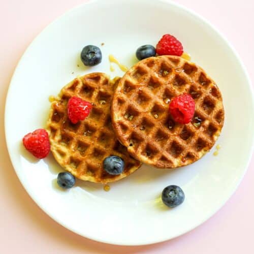 Two golden oat flour waffles are served on a white plate with fresh raspberries and blueberries, drizzled with syrup on top.