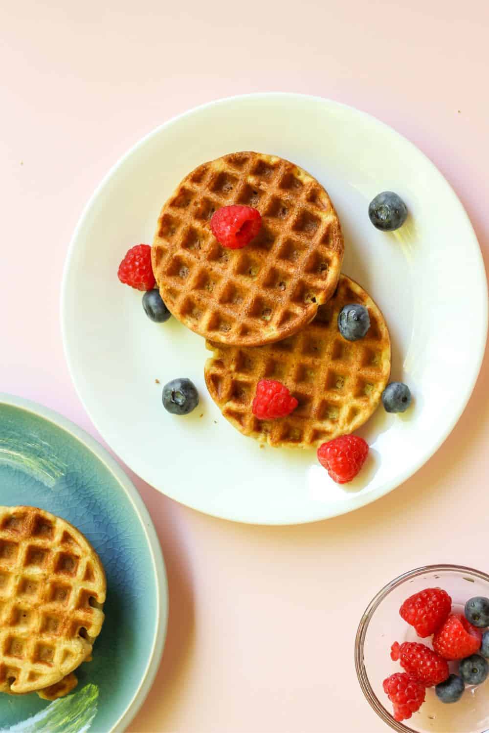 Overhead view of a white plate with two waffles topped with fresh raspberries and blueberries. There is one-fourth of a blue plate with a waffle on the left bottom side.