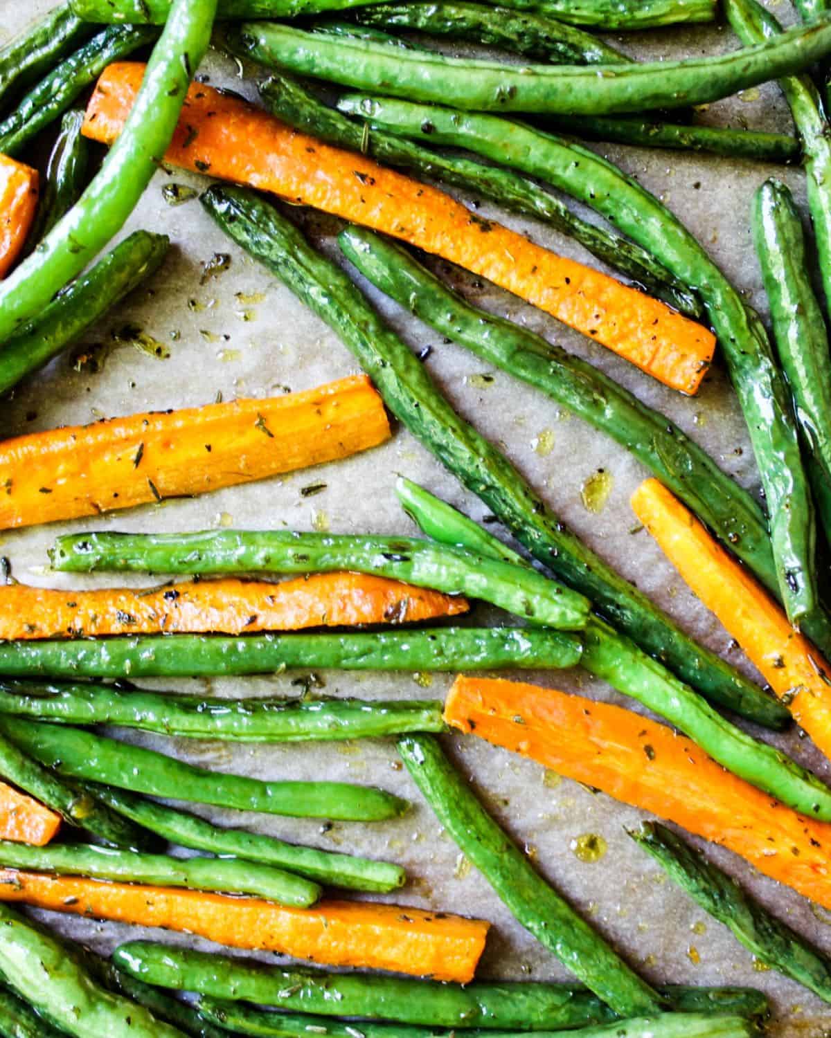 Roasted carrot sticks and green beans with dry herbs and coated in oil on yellow parchment paper.