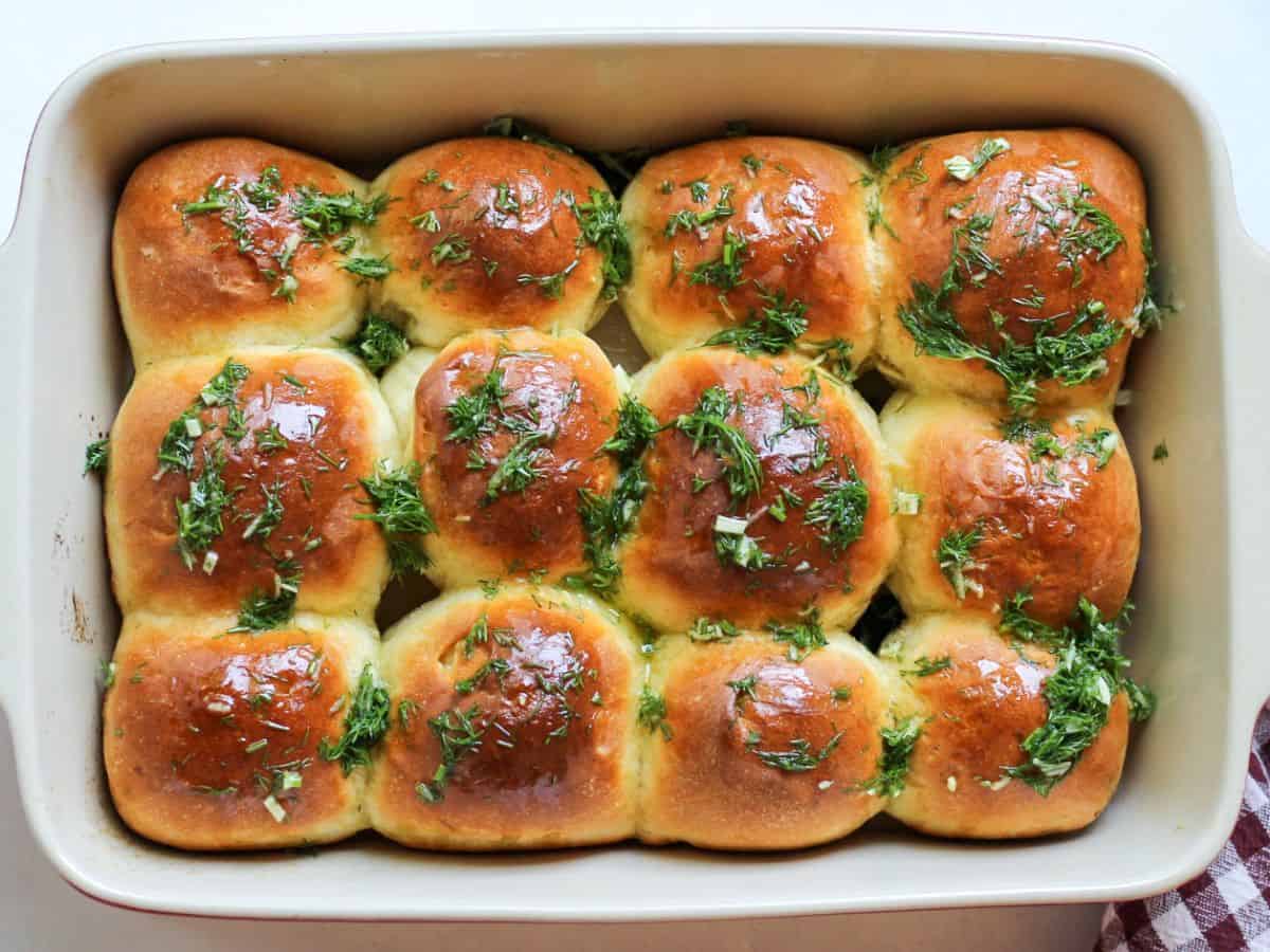 A rectangular baking ceramic dish with 12 baked bread rolls topped with chopped green herbs.