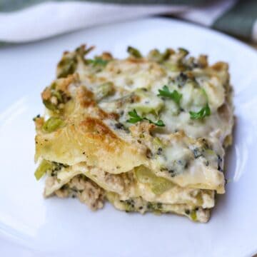 A square cut of cooked white lasagna on a white plate. There are with few green parsley leaves on top.