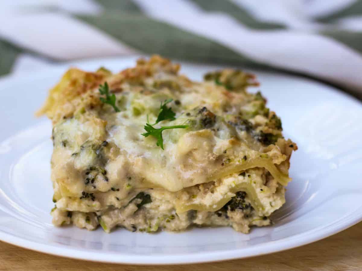 A square cut of cooked white lasagna on a white plate. There are with few green parsley leaves on top and a green-white material in the background.