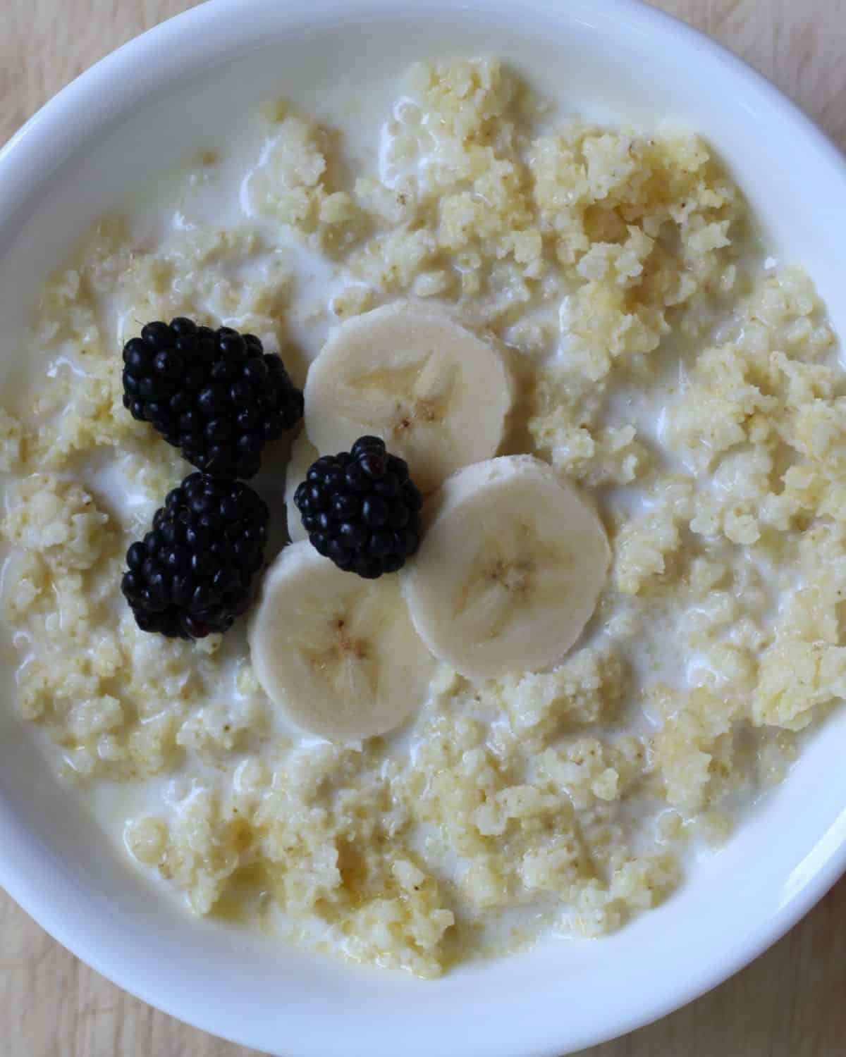 Yellow grain porridge with milk topped with slices bananas and fresh blackberries in a white bowl.