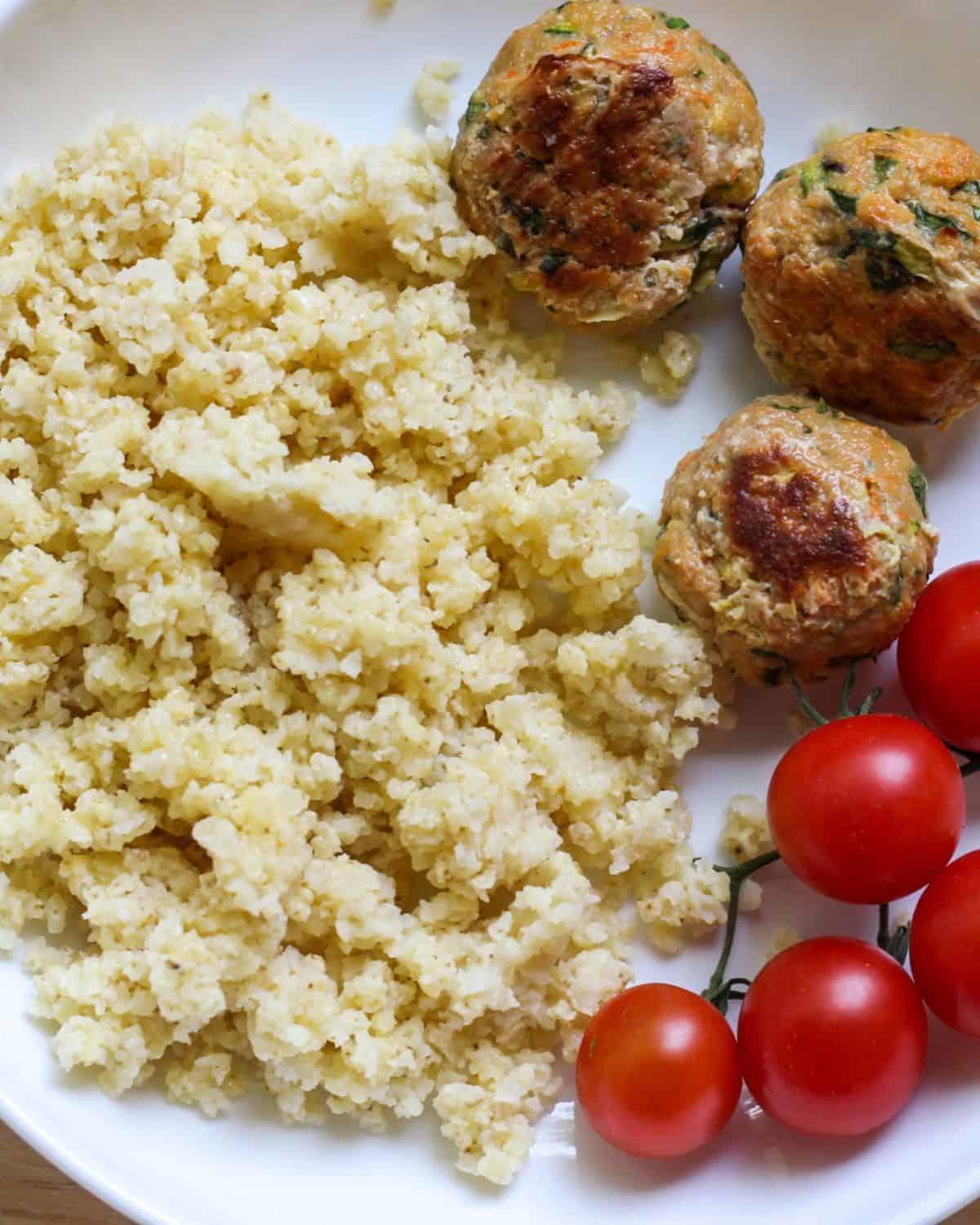 Cooked yellow grain, three meatballs and red cherries tomatoes on a vine served on a white plate.
