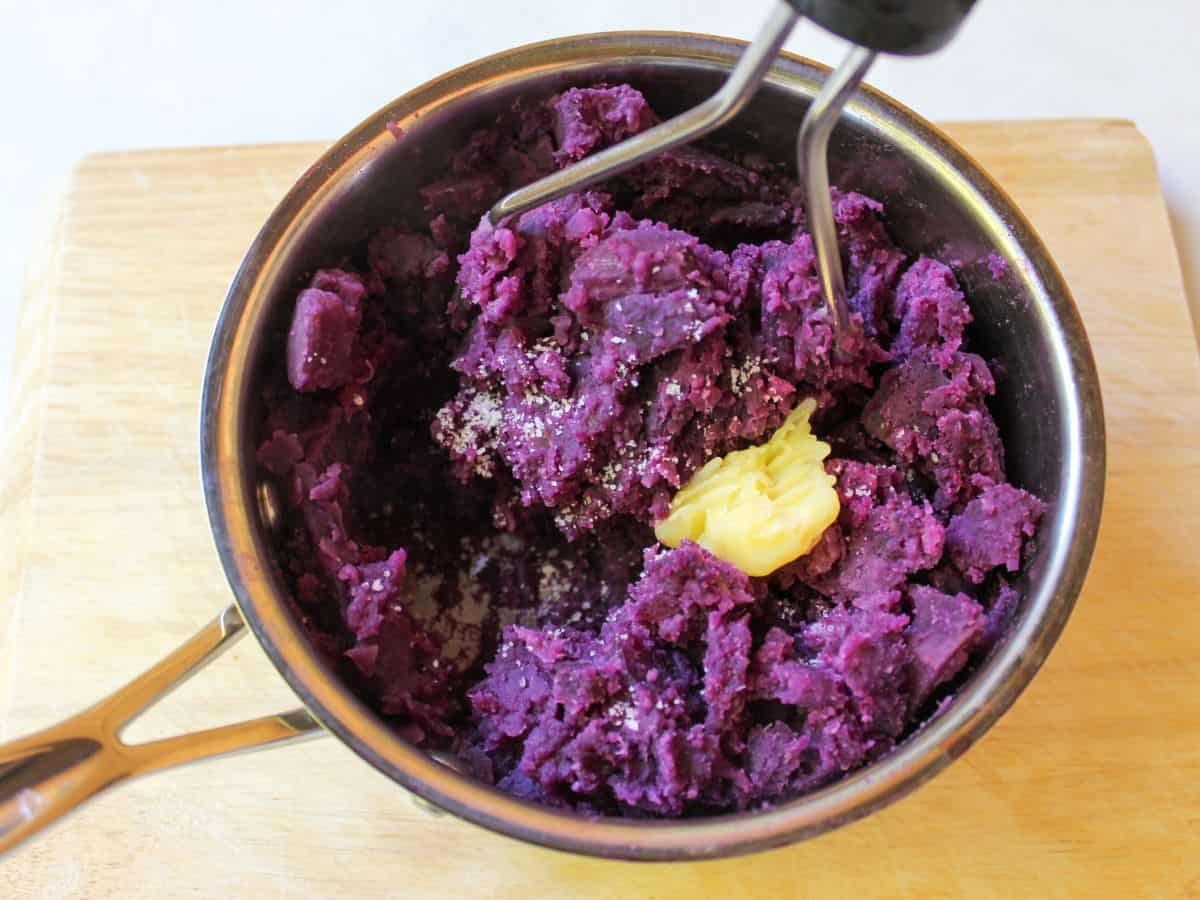 A stainless steel pot with mashed purple puree, a potato masher, salt crystals and a scoop of ghee butter on top.