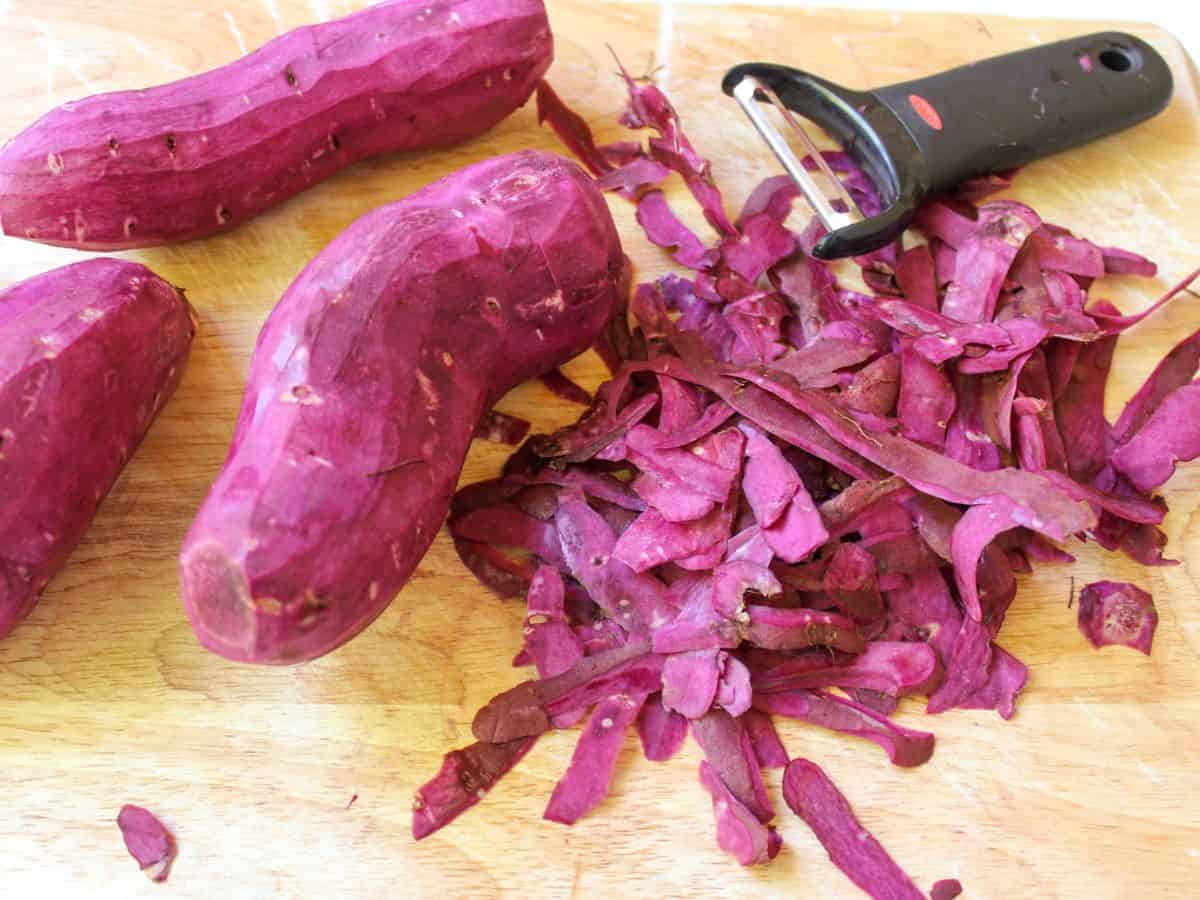 A cutting board with 2 long purple potatoes, a veggie peeler, and a pile of loose peels.