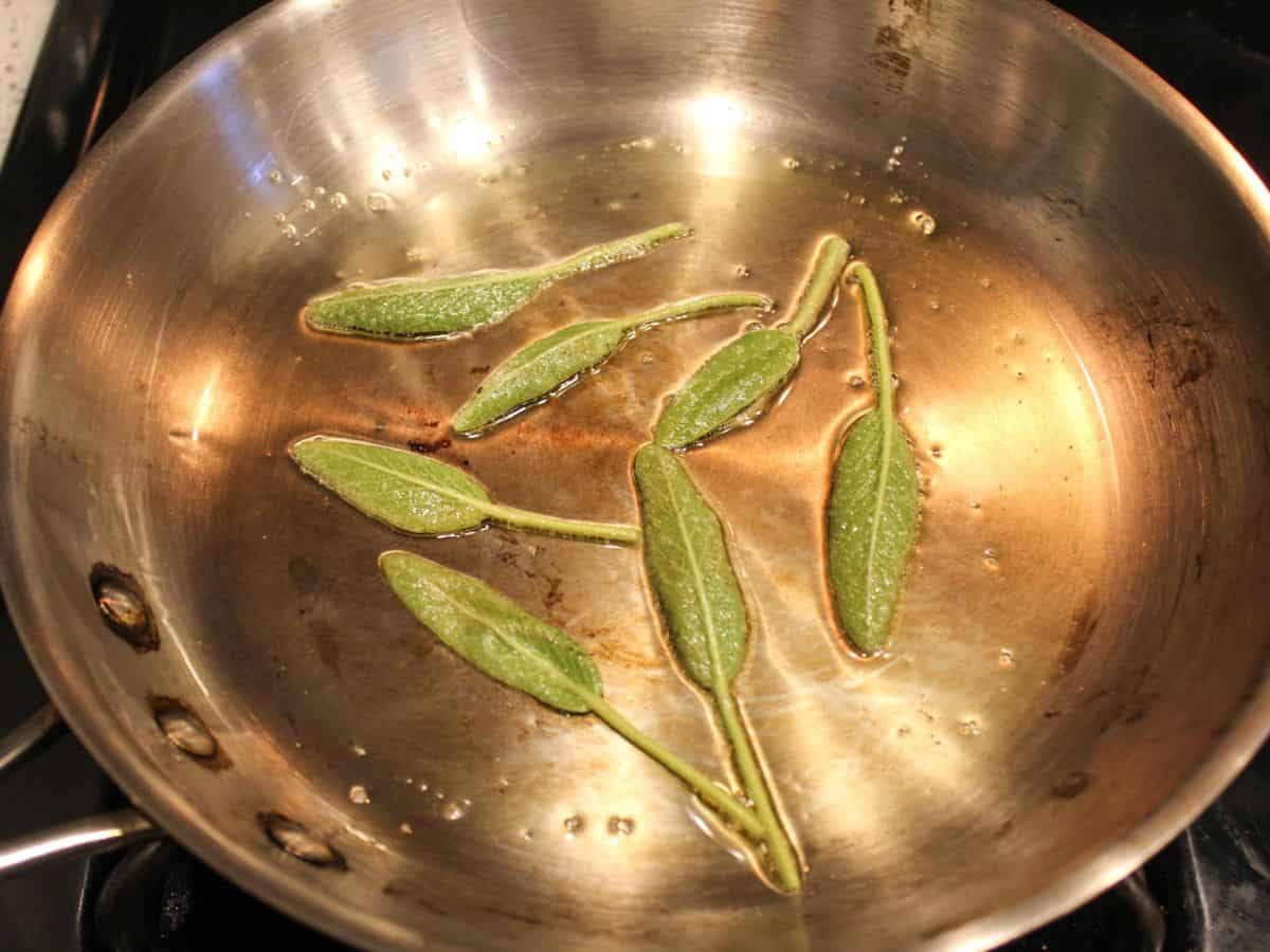 Stainless steel skillet with oil and seven whole green sage leaves.