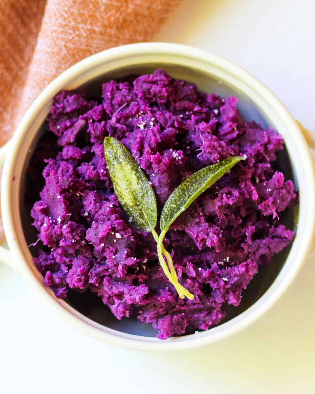 Overhead view of white bowl with bright purple puree, large crystals of salt, and two whole sage leaves on top. The backrgound is white with some bage-brown cloth material in the top left corner.