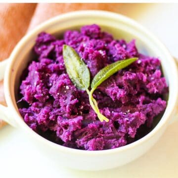 A white bowl with bright purple puree, large crystals of salt, and two whole sage leaves on top. The backrgound is white with some bage-brown cloth material in the top left corner.