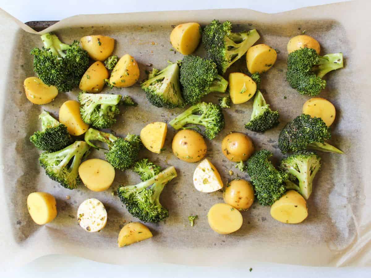Raw yellow halved potatoes and green broccoli are arranged on a large baking pan covered with parchment paper..
