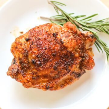 A turkey thigh with crispy and golden skin on a white plate. There is some fresh green rosemary on the plate.