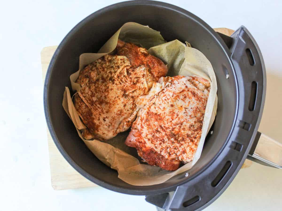 Air fryer basket covered with parchment paper with two raw seasoned thighs.