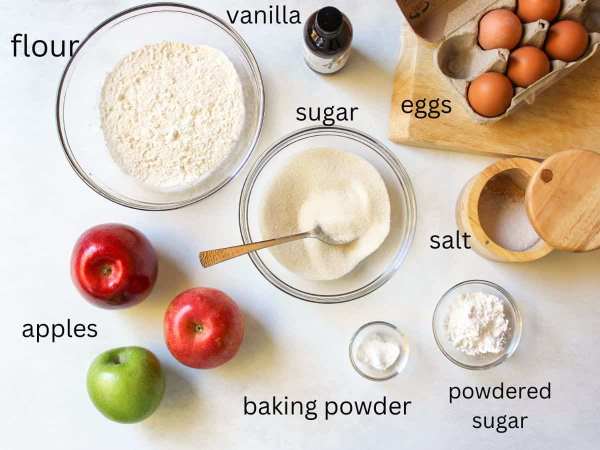 Sponge apple cake recipe ingredients on a white surface. They are labeled with corresponding names.