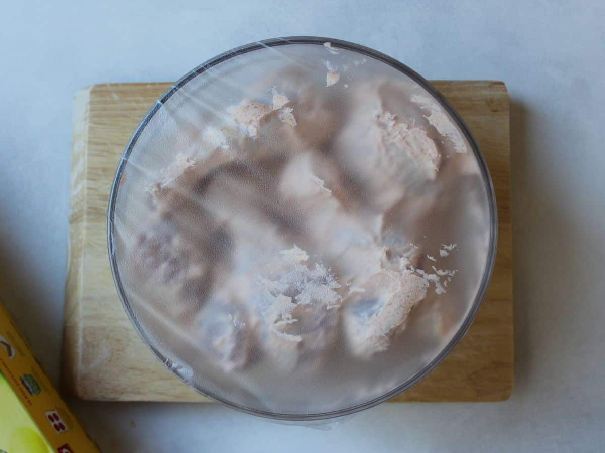 A glass bowl with raw chicken legs in madinade covered with some plastic clear wrap.