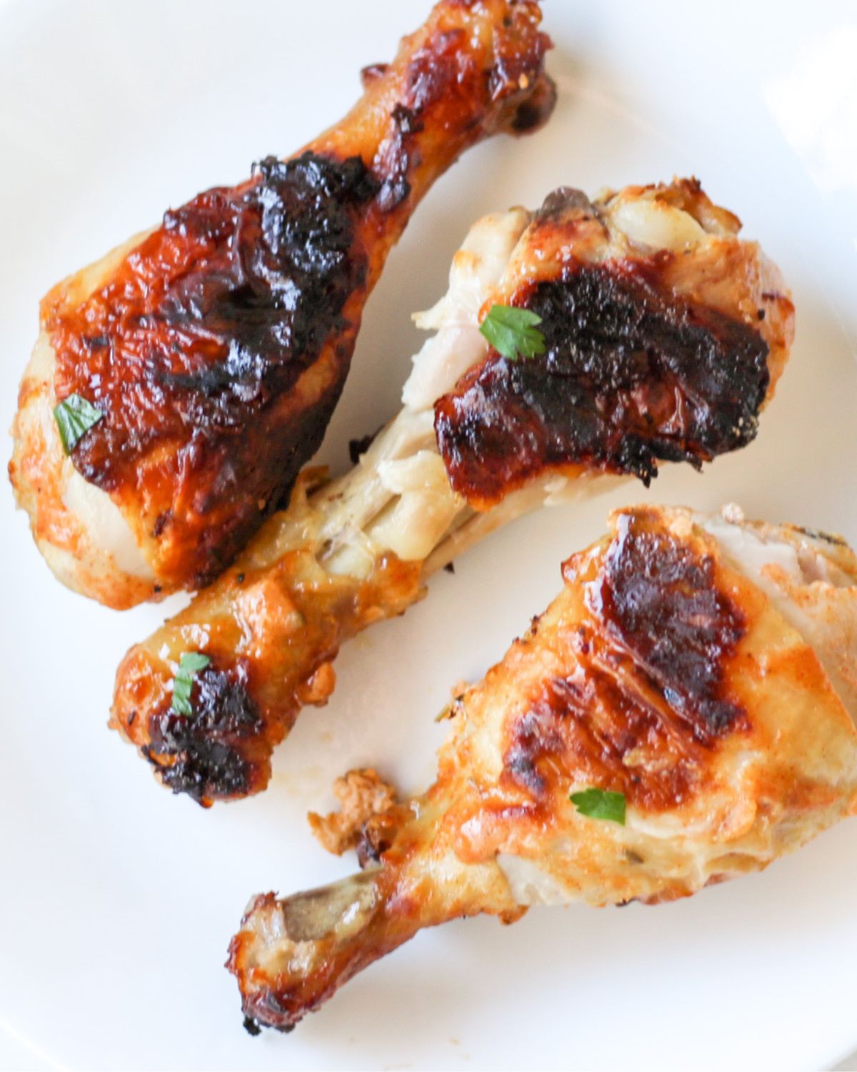 Three cooked chicken drumsticks on a white plate topped with small pieces of green parsley..