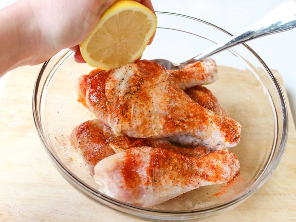 A glass bowl with raw chicken legs coated in red seasoning. The lemon is being squeezed over the chicken.