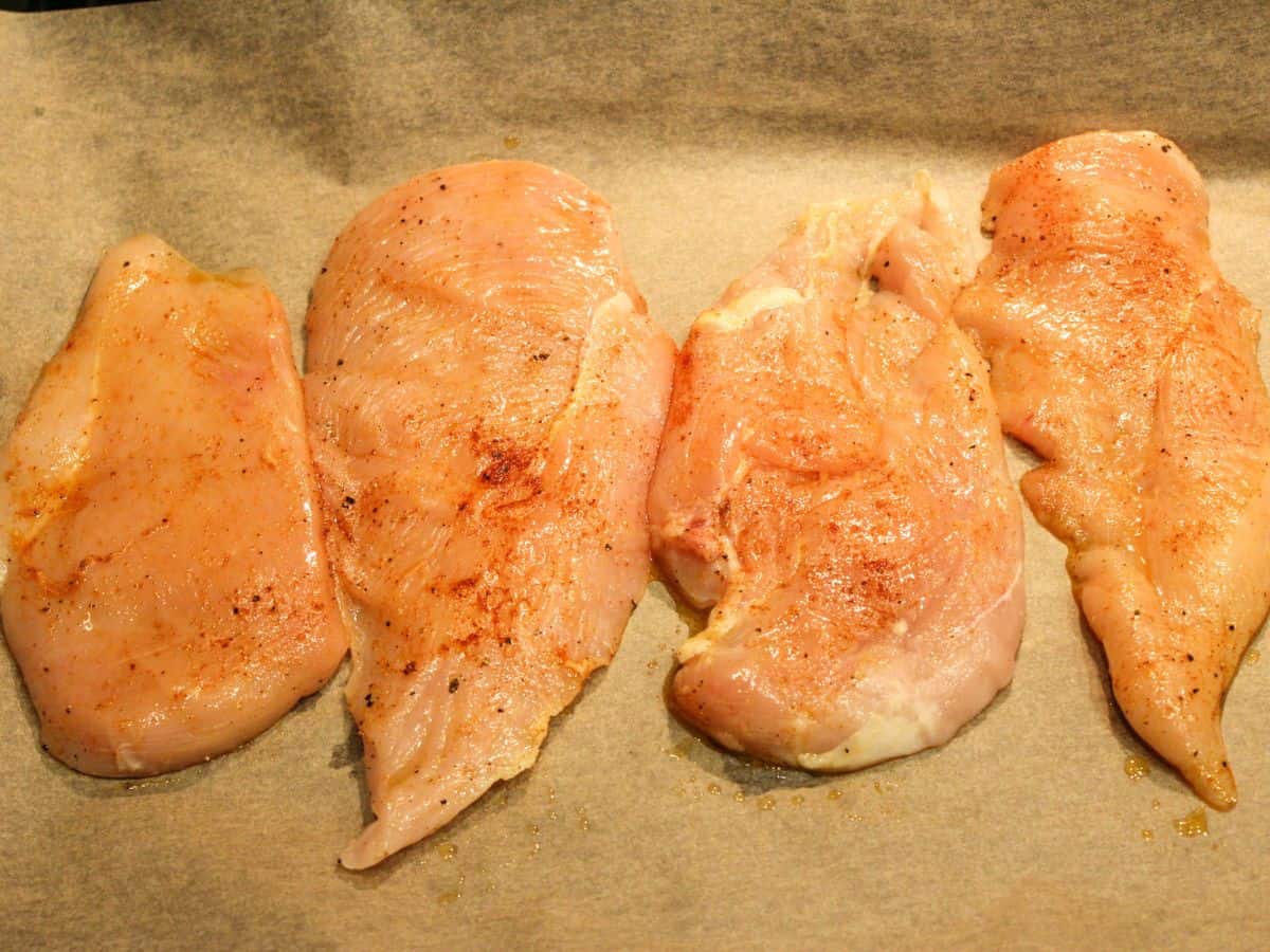 4 seasoned raw chicken breasts on a parchment paper arranged vertically.