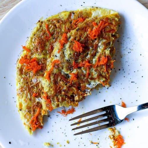 Folded in half omelette with freshly grated carrots on top and some black pepper. There is a fork on the plate.