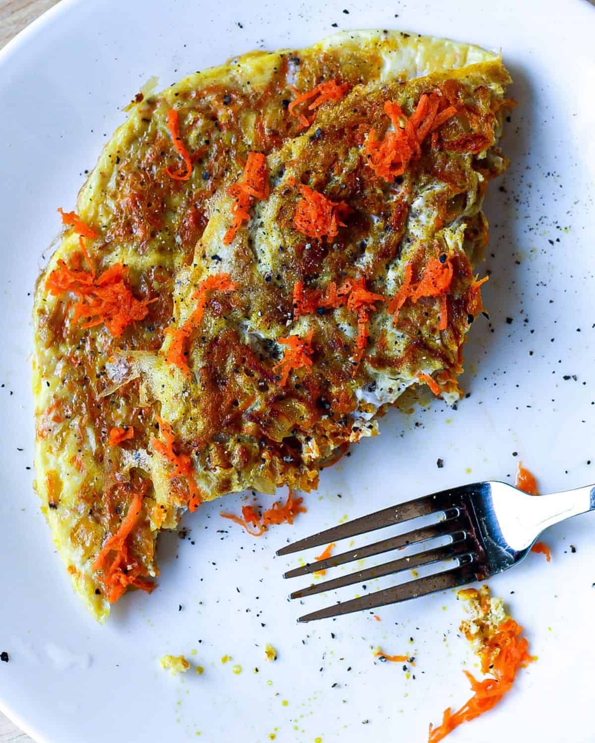 Folded in half omelette with freshly grated carrots on top and some black pepper. There is a fork on the plate. 